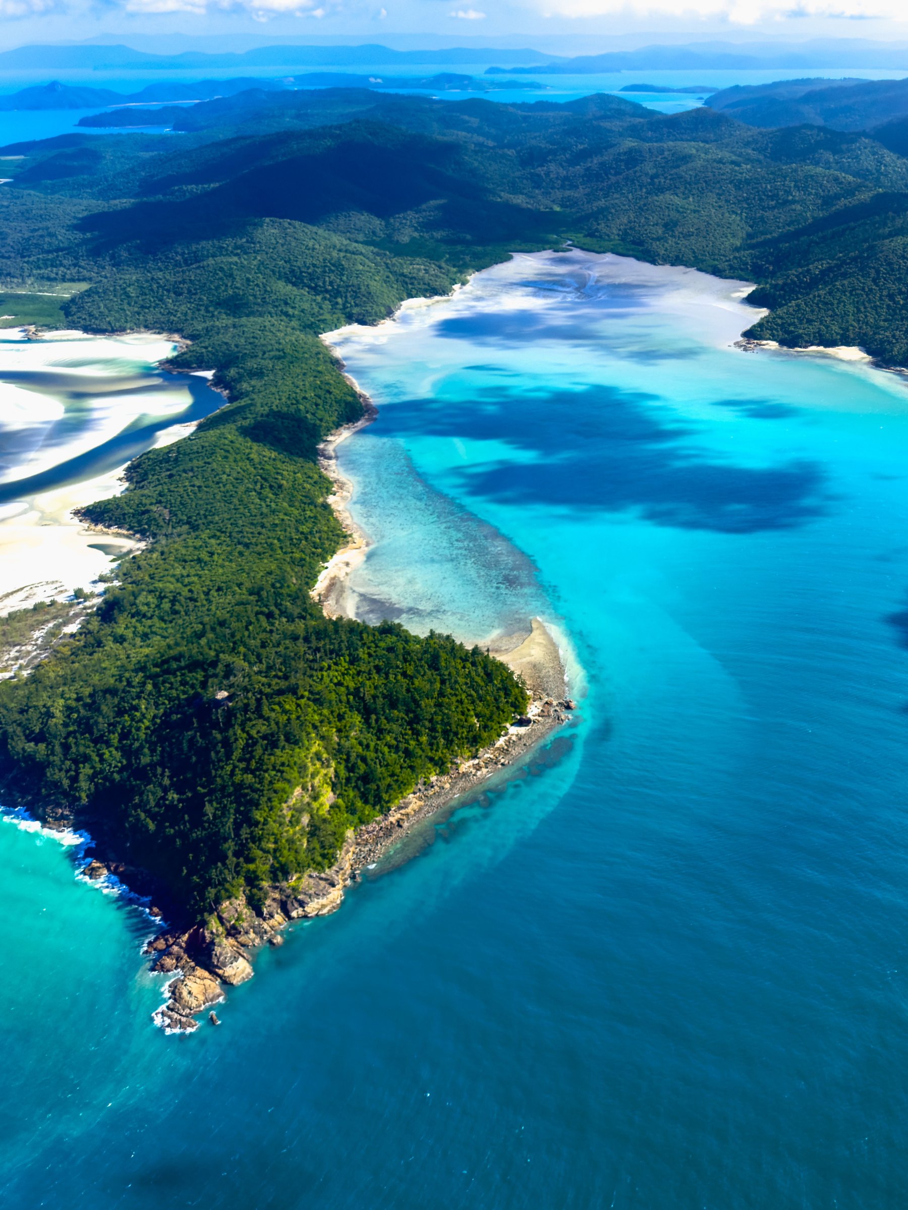 A forested Whitsunday island ringed by white sand and turquoise water, seen from the air